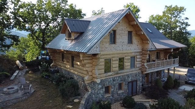 Two-story log cabin with stone foundation and metal roof, surrounded by trees.