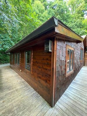 Wooden cabin with dark brown siding and windows, on a wooden deck surrounded by trees.
