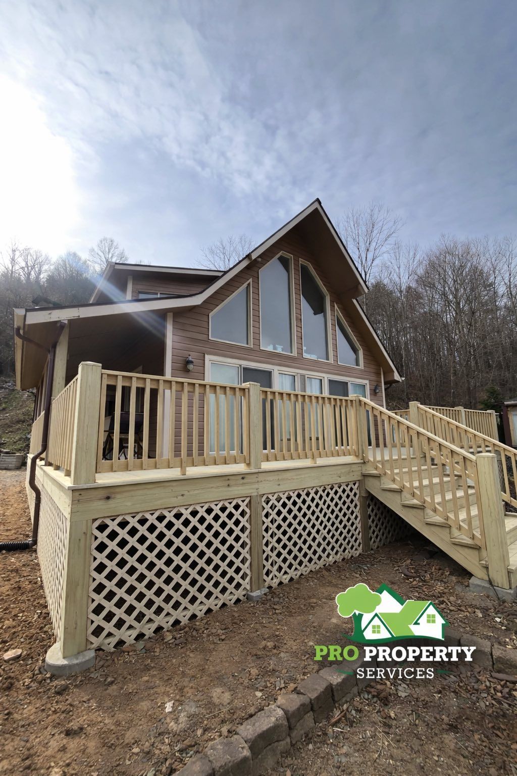 A two-story house with a wooden deck and lattice. The house has large front windows and is surrounded by bare trees under a cloudy sky.