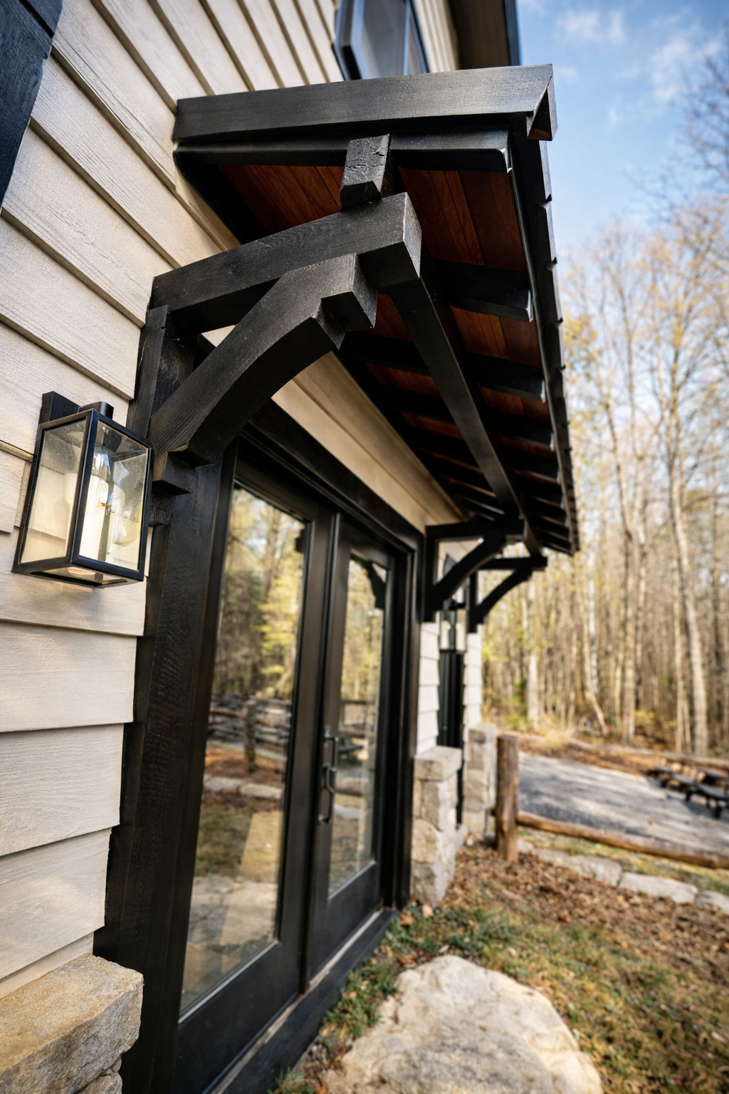 Black wooden awning over glass doors, on a light gray-sided building with stone accents; forest background.