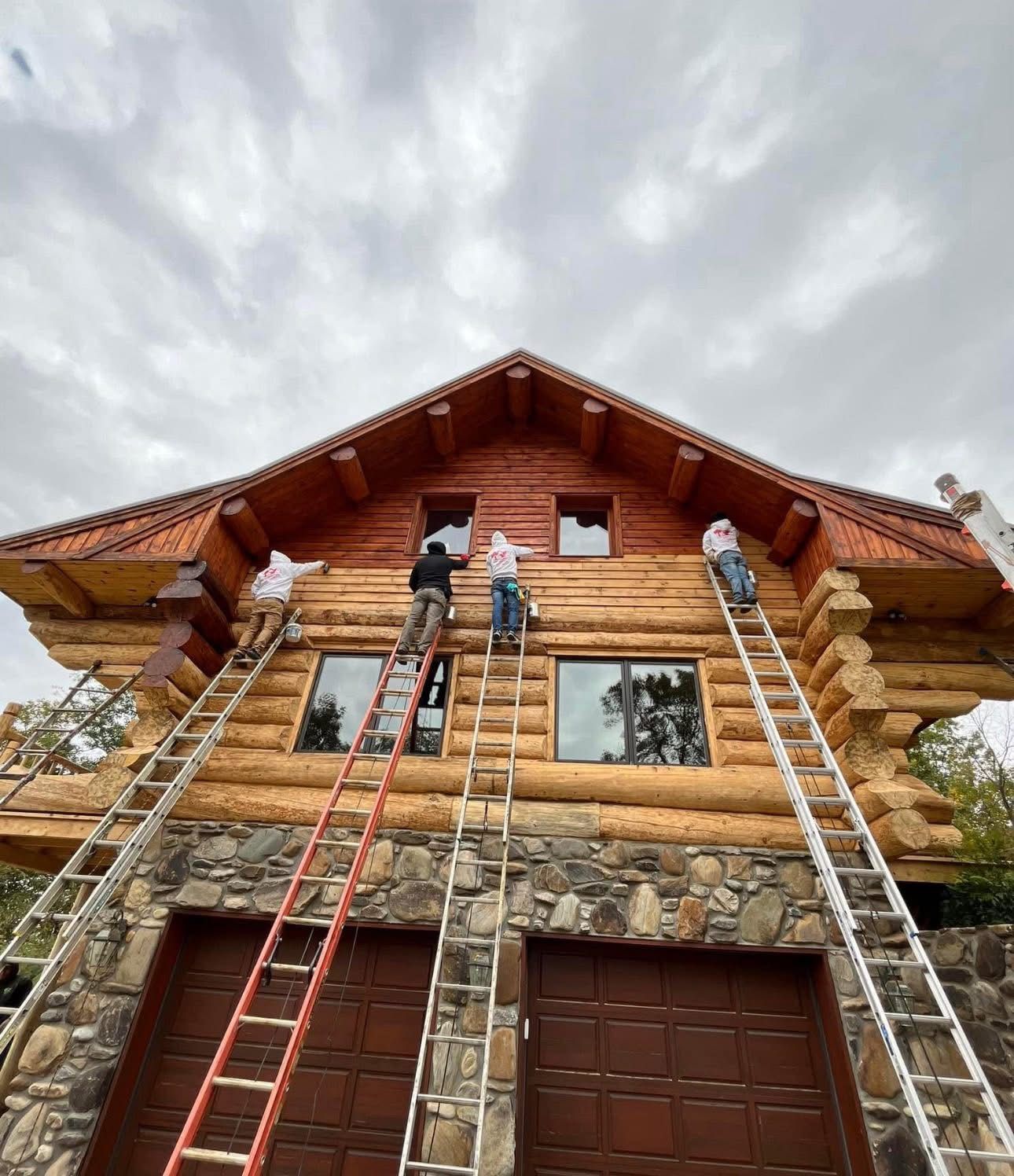 Four people on ladders painting the wooden exterior of a two-story log cabin under a cloudy sky.
