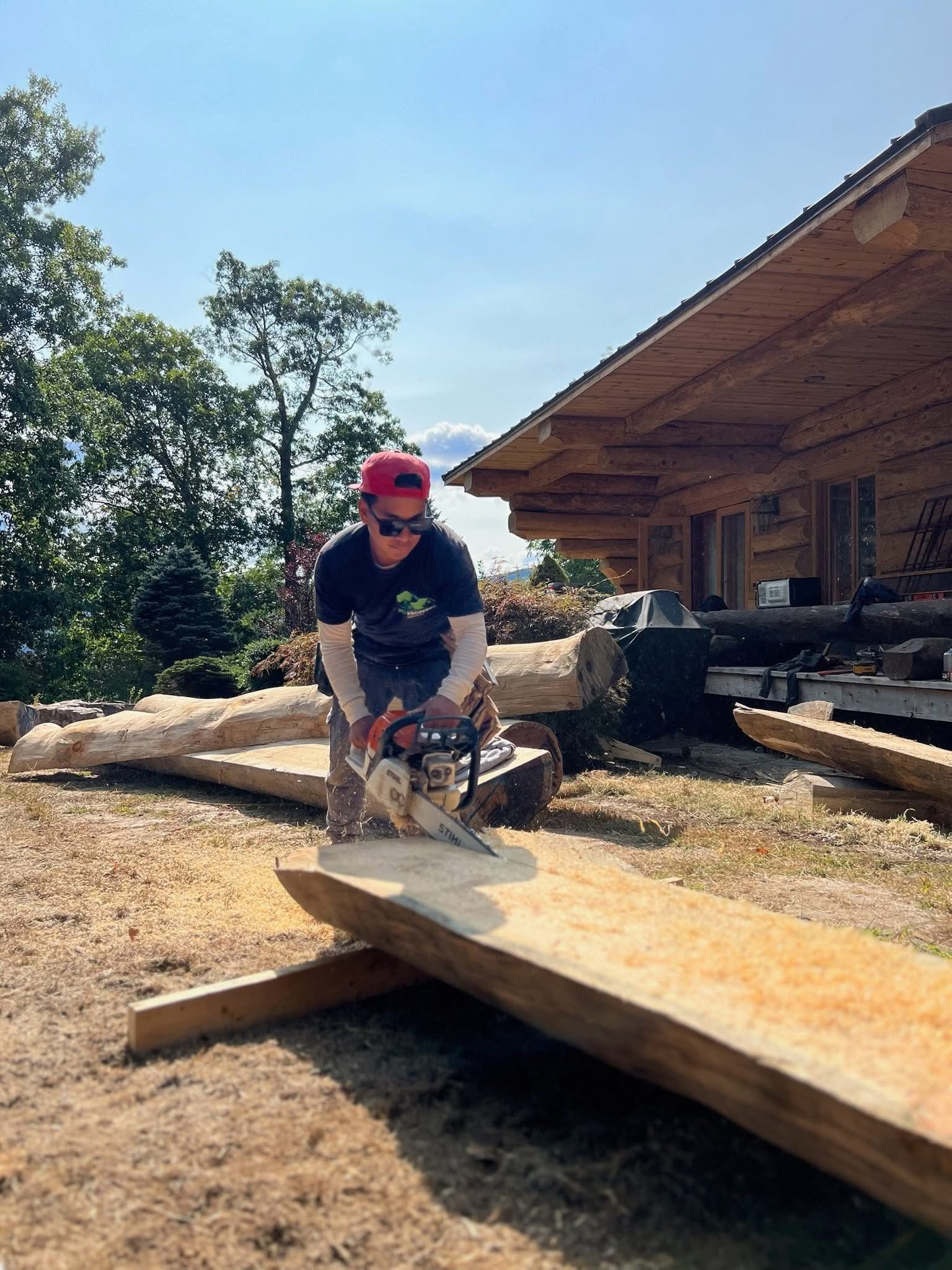 Person using a chainsaw to cut a wooden plank outdoors. Log cabin visible in the background. Sunny day.