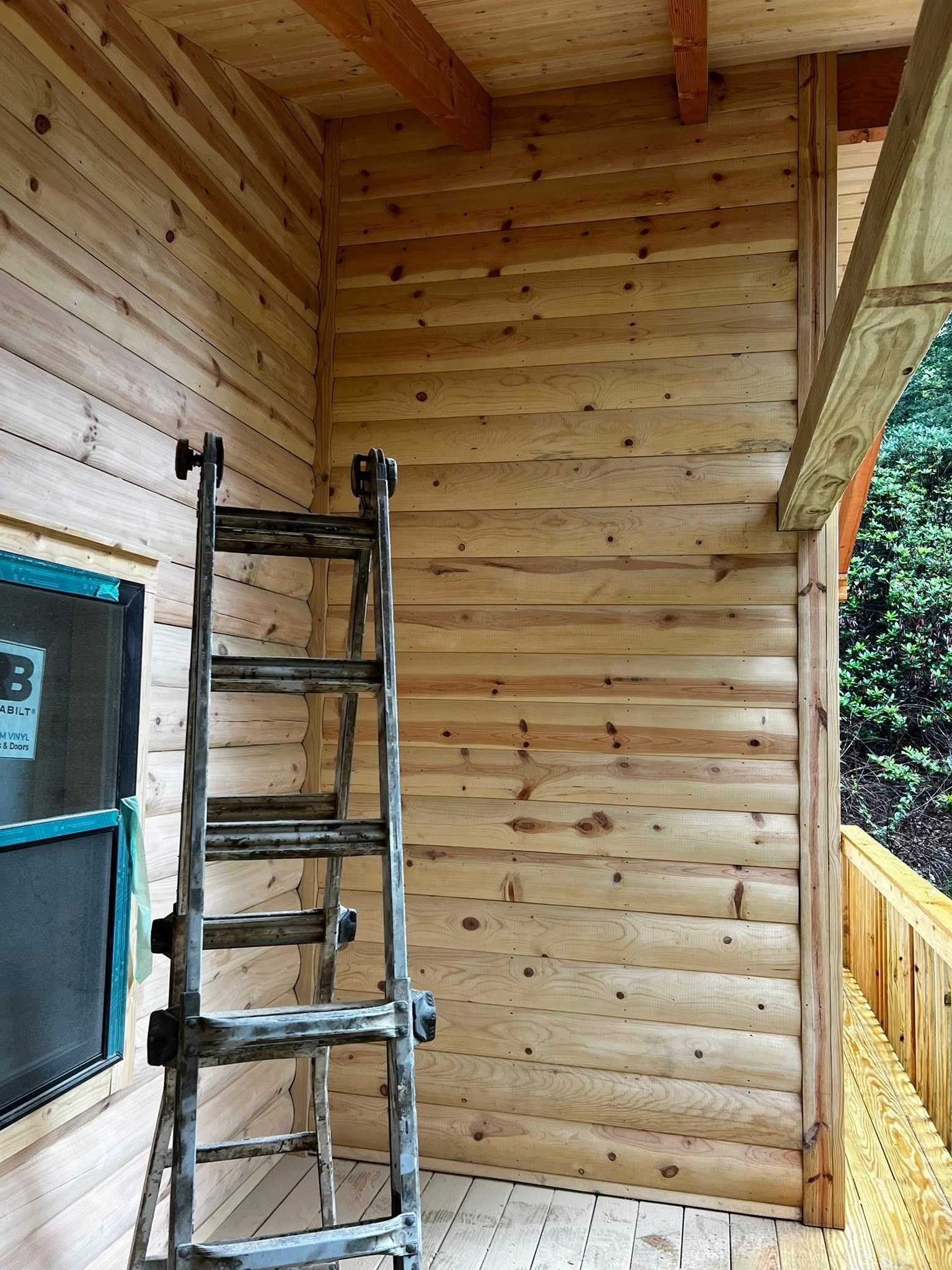 A person in red cap using a chainsaw to cut wood outdoors near a log cabin.