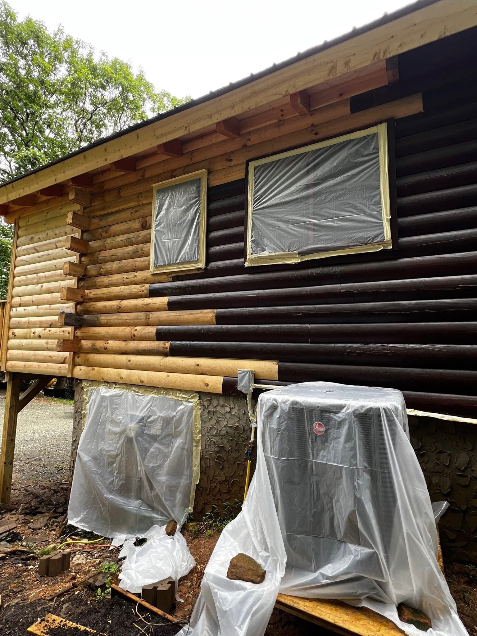 A log cabin being stained. Half-painted with a dark brown stain, windows covered with plastic.
