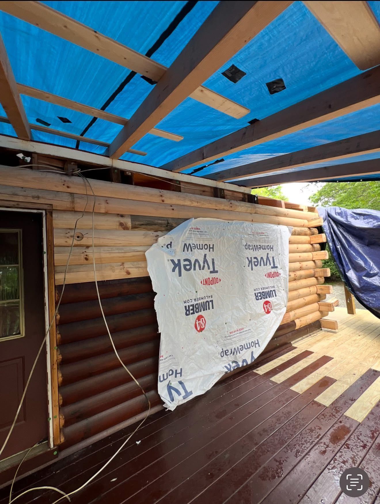 Log cabin exterior under construction, covered with blue tarp. Tyvek wrap partially covers the logs.