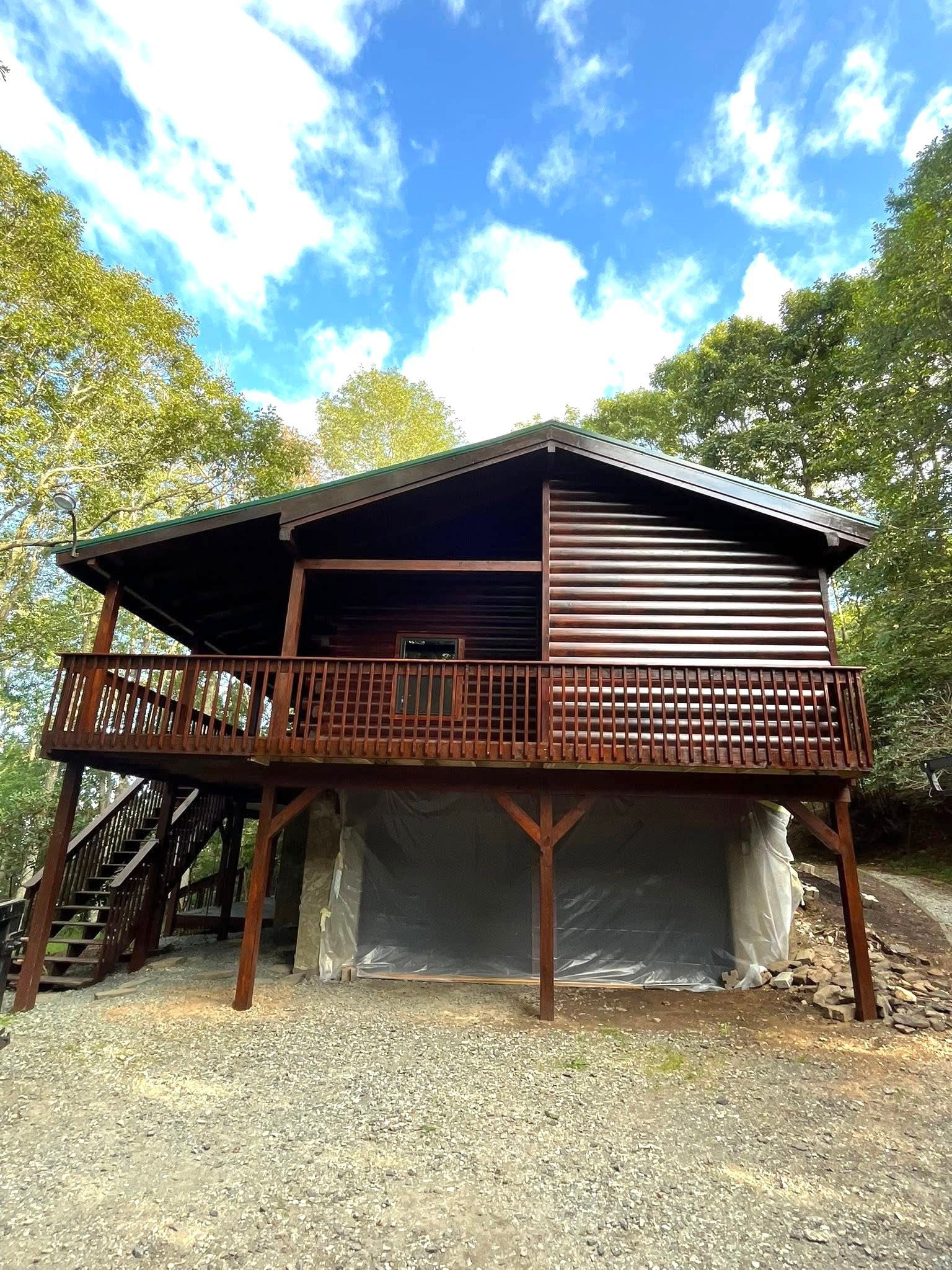 Wooden cabin with a deck, surrounded by trees under a blue sky.