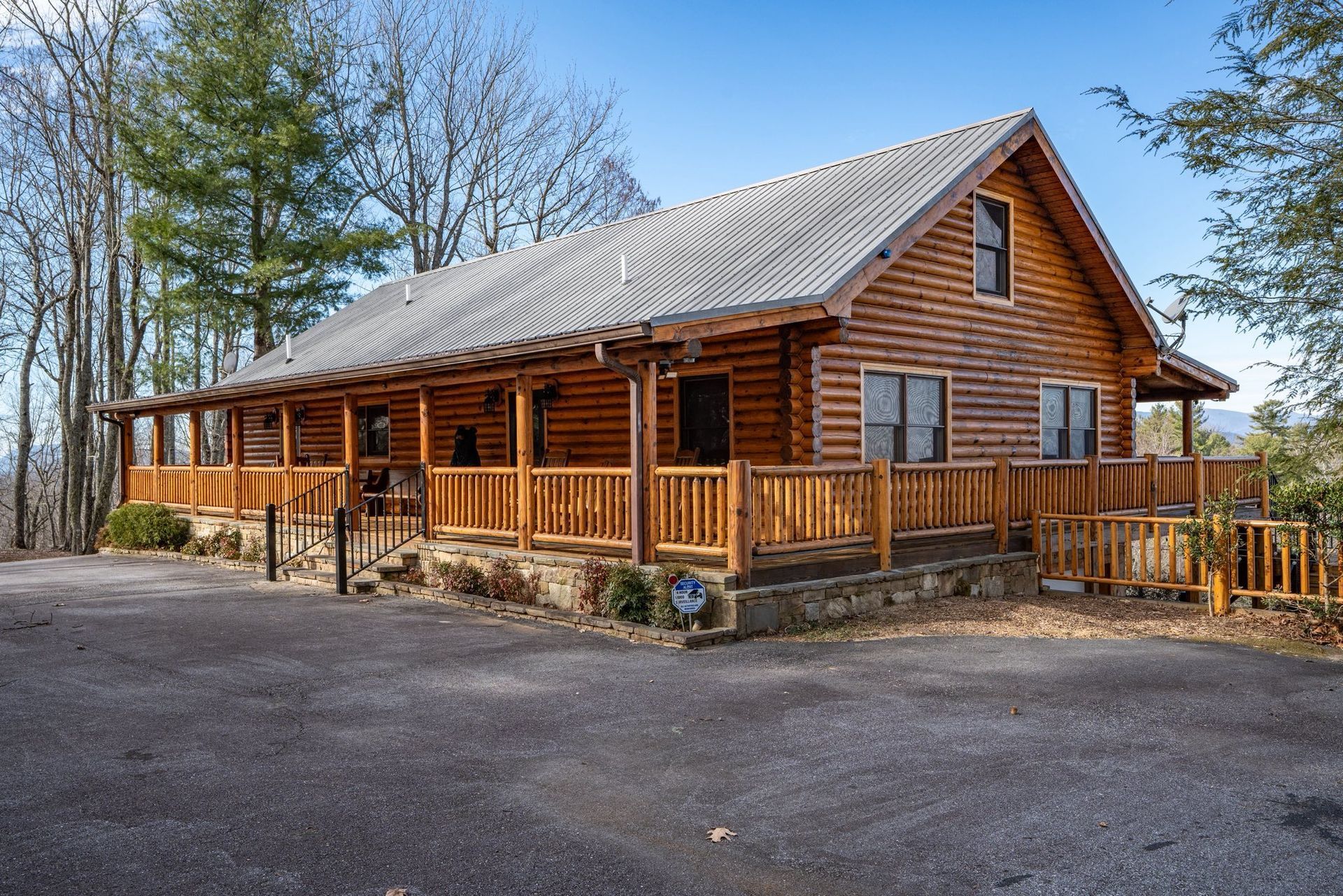Wooden cabin with a wrap-around porch, on a hill, surrounded by trees.
