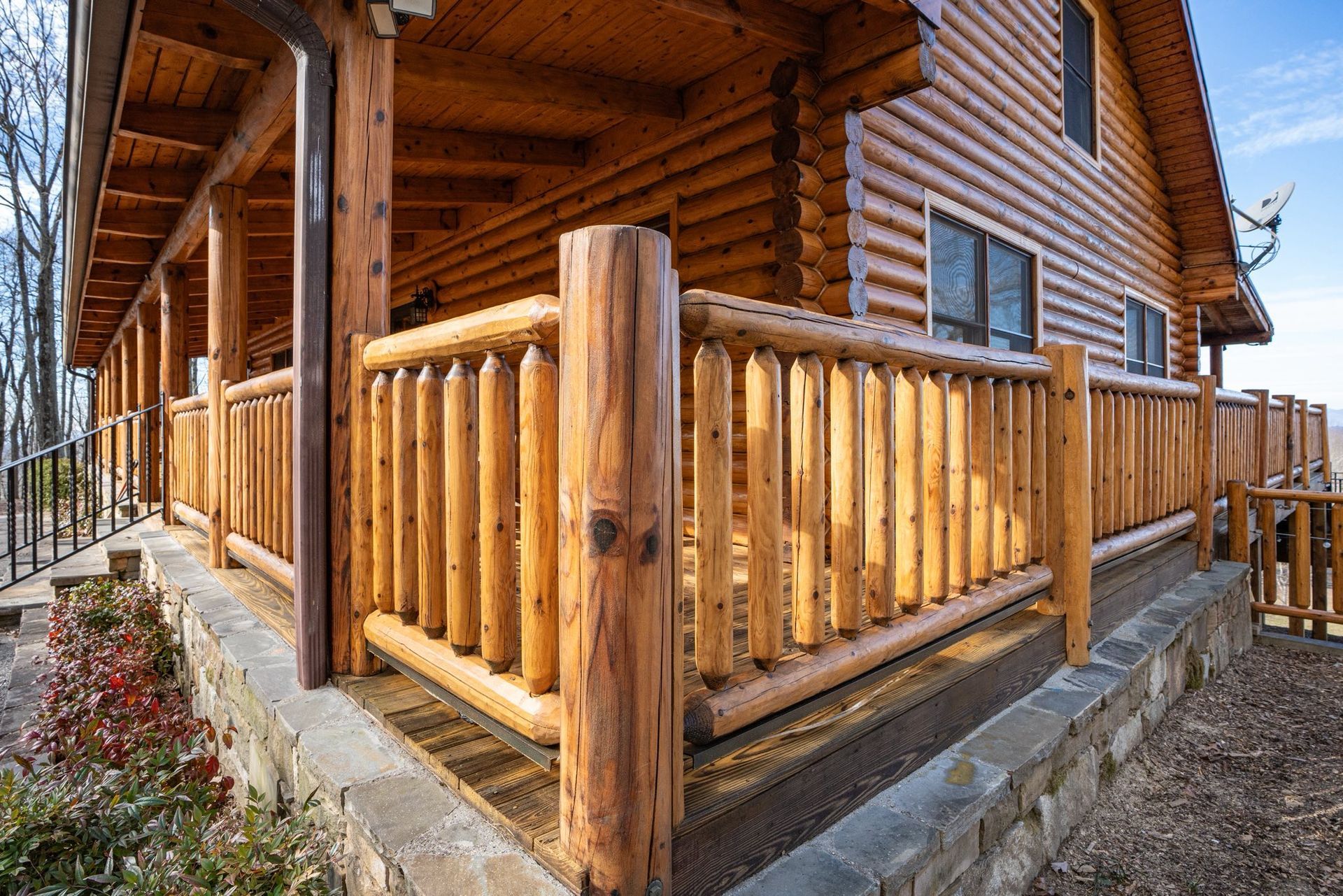 Log cabin with wooden railings on a porch, surrounded by a stone base and trees against a blue sky.