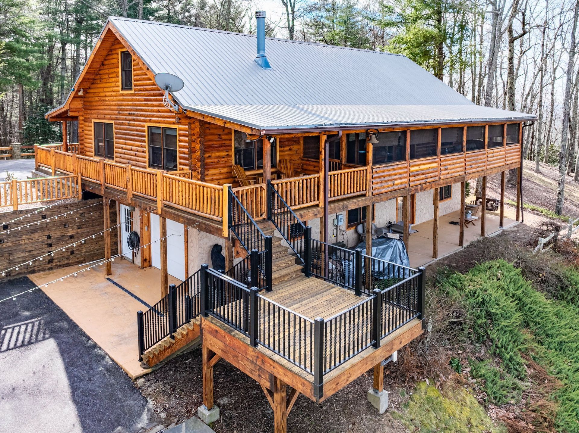 Log cabin with wraparound porch and deck, nestled in wooded area. Exterior is wood with a silver roof.