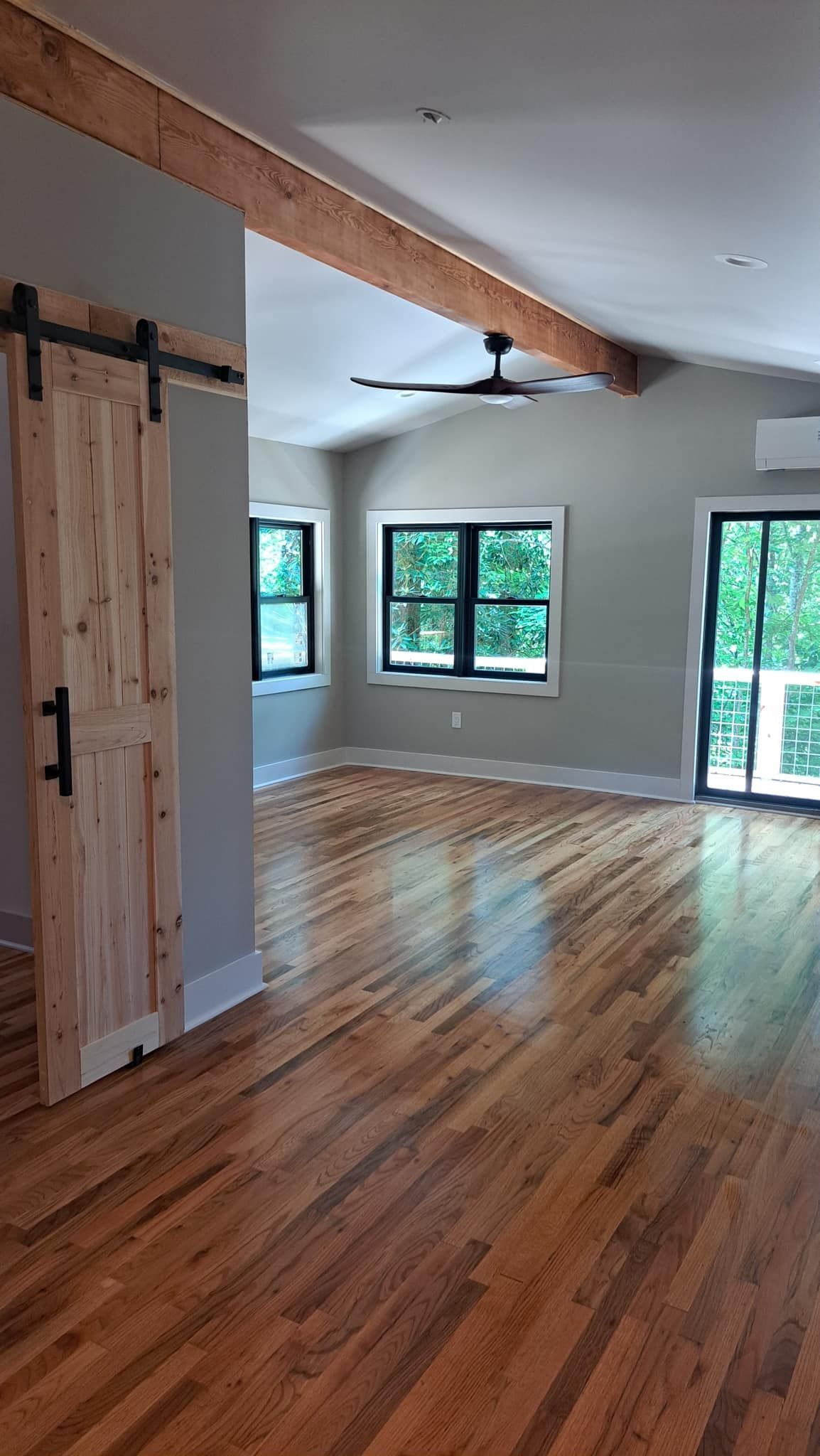Wooden interior with sliding barn door, exposed beams, hardwood floors, windows, and a ceiling fan.