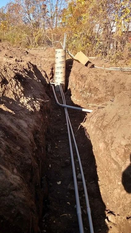 Trench in dirt with gray pipes leading to a concrete-like pillar; sunny outdoor setting.