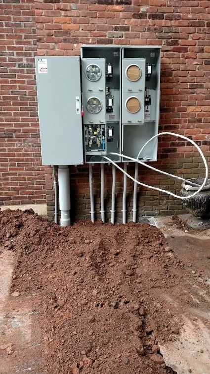 Electrical meter boxes mounted on a brick wall with conduits, wires, and a pile of dirt in the foreground.