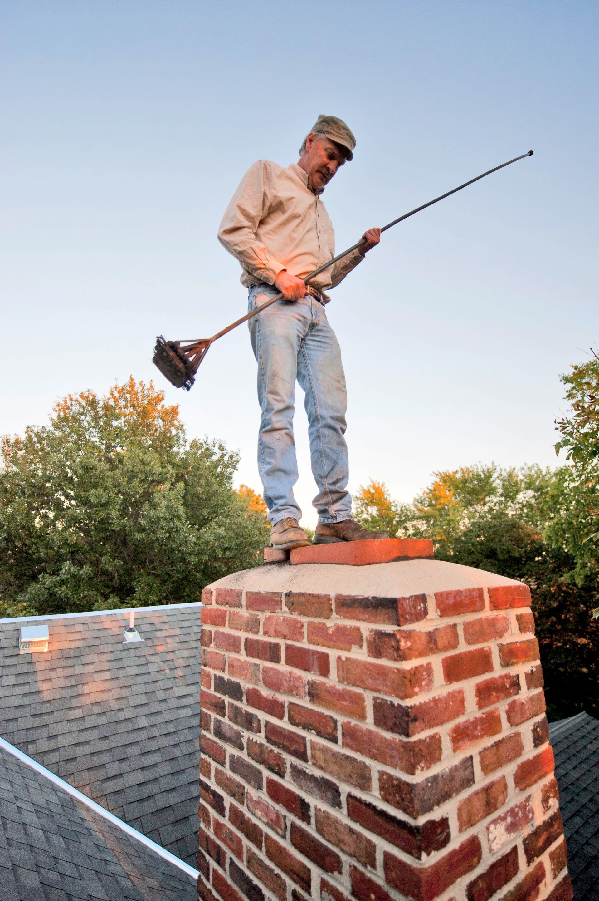 Man on a chimney sweeping it with a long brush. Sky is clear. Brick chimney against a roof.