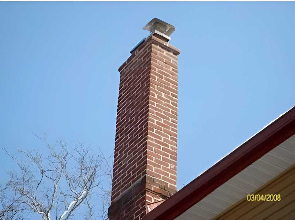 Tall brick chimney with a metal cap against a blue sky, next to a house's roof.