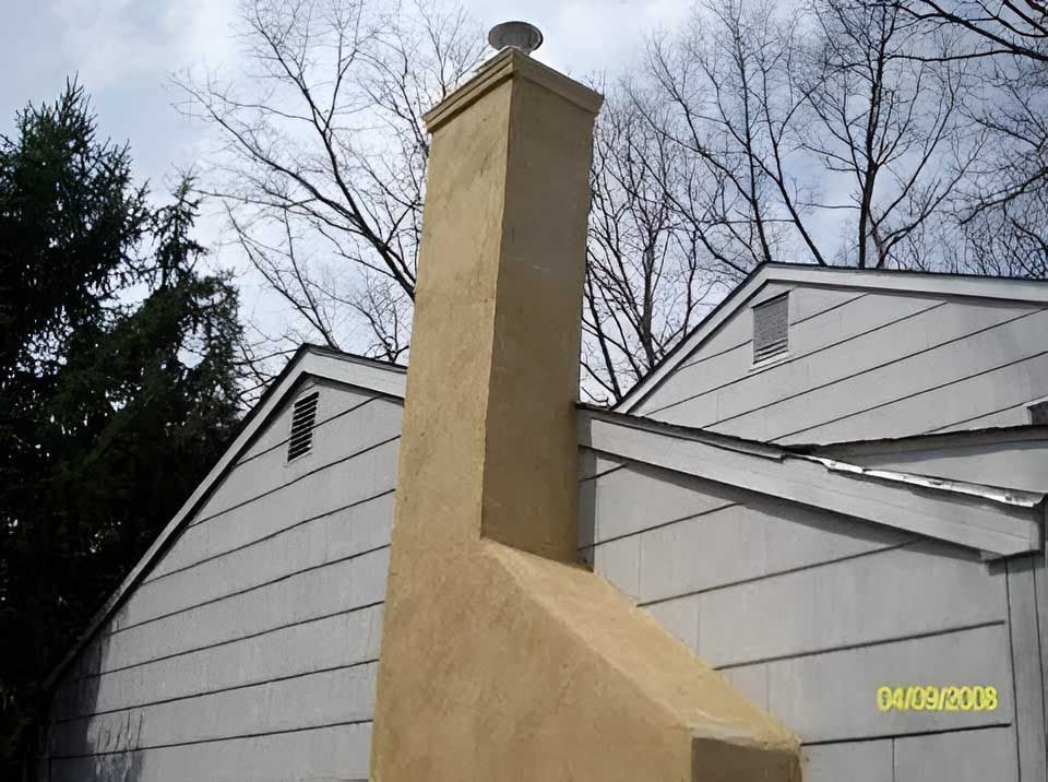 Tan stucco chimney on a white-sided house, against a sky with leafless trees.