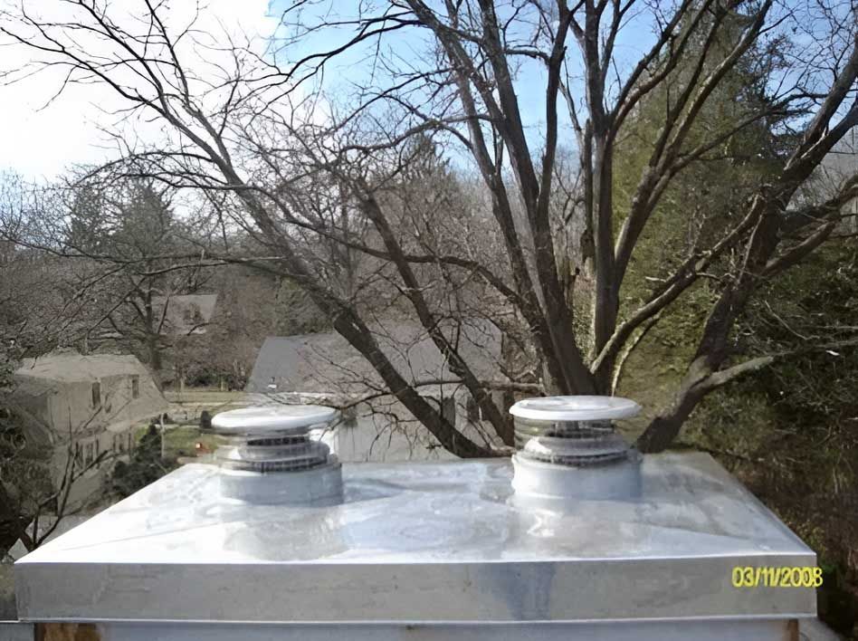 Two metal chimney caps with spinning vents on a roof, with trees and buildings in the background.