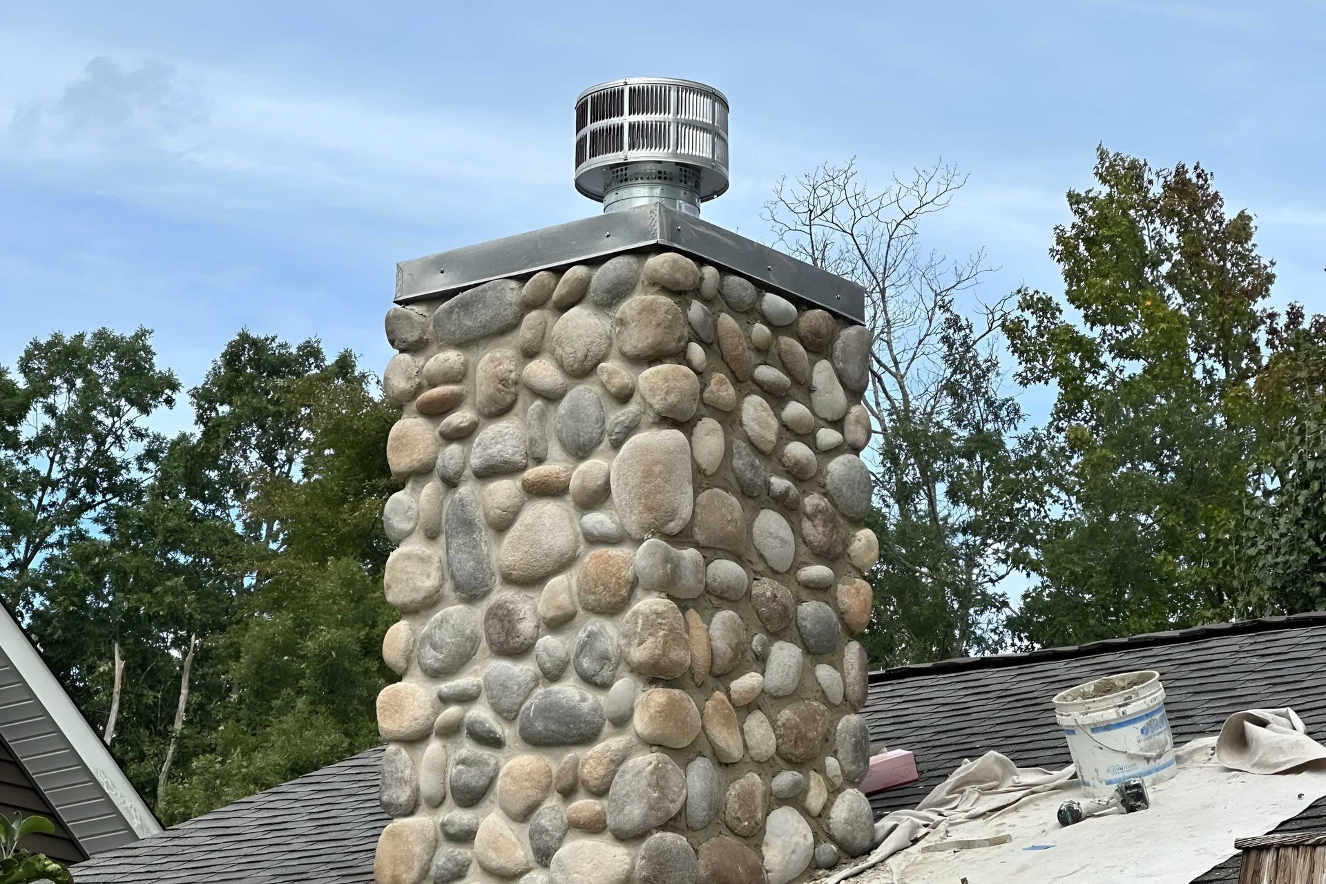 Stone chimney with metal cap on a rooftop, surrounded by trees.