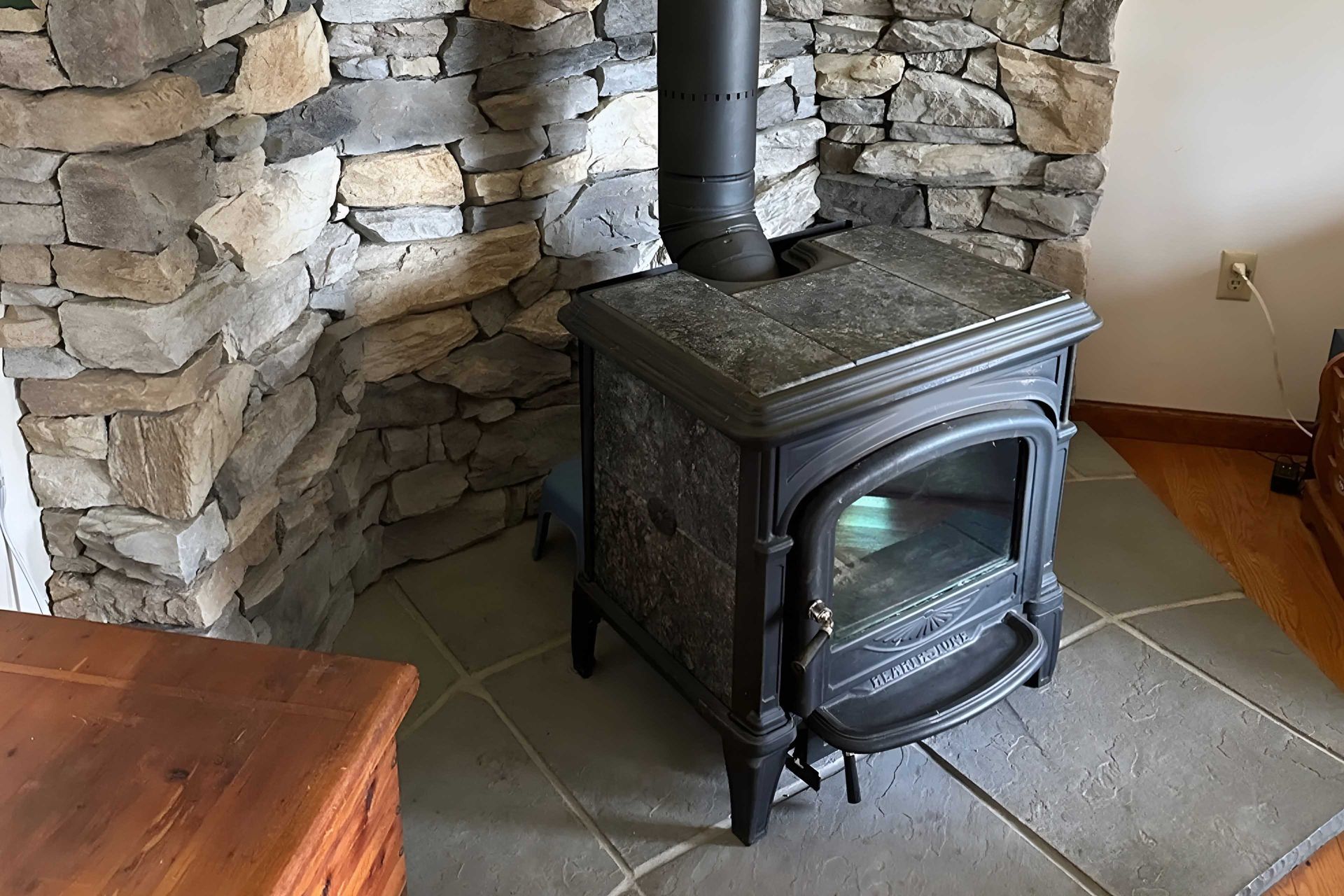 Wood-burning stove with black chimney pipe, set against a stone wall, on a tiled floor.