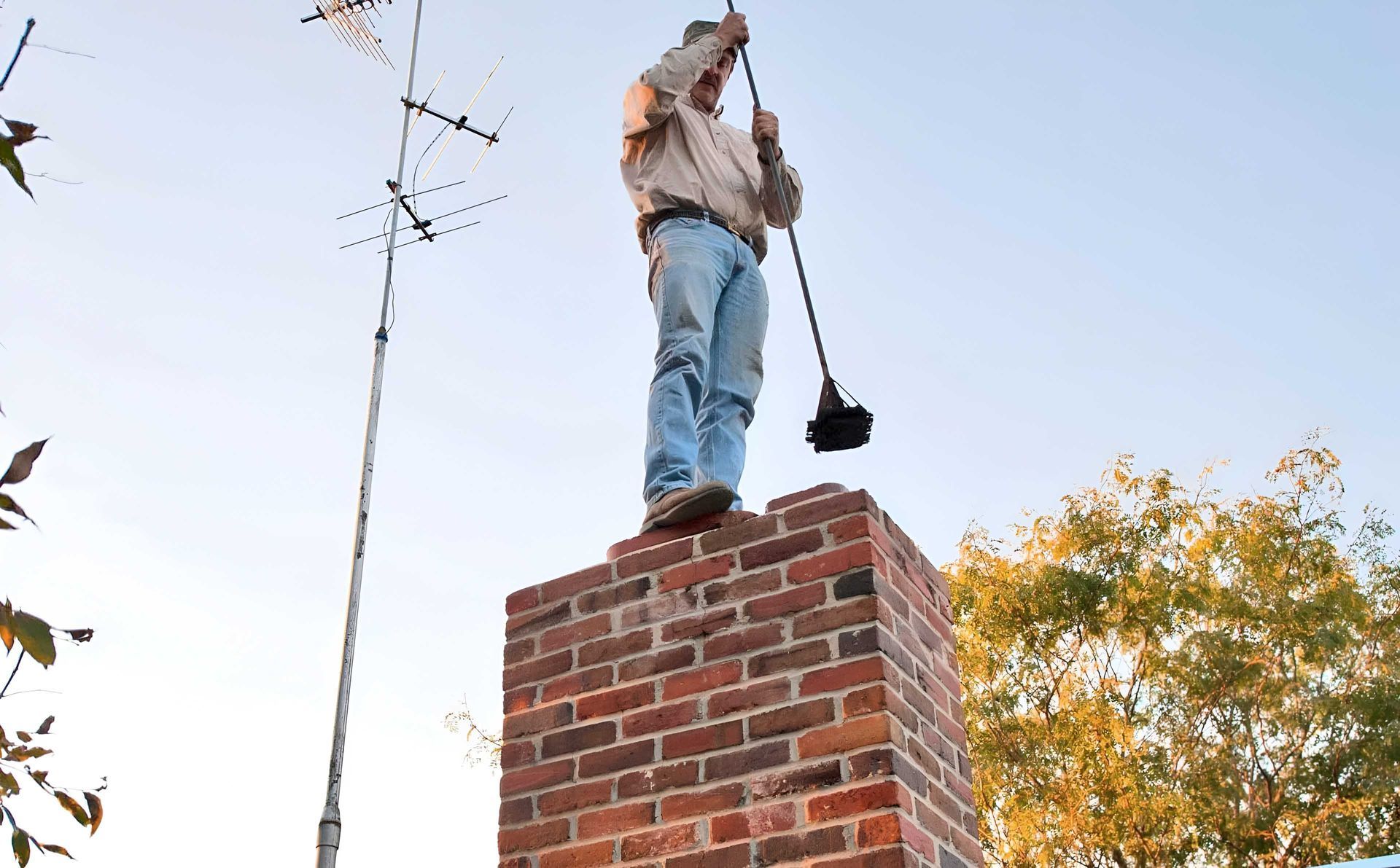 Man cleaning a chimney with a brush while standing on top of it; blue sky in the background.