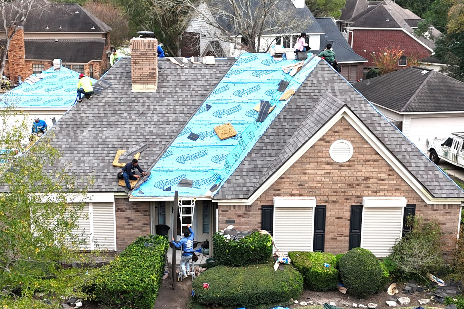 Roofers replacing a shingle roof on a brick house, blue underlayment visible.