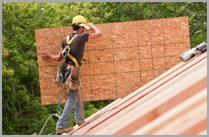A man is standing on a roof holding a large piece of plywood.