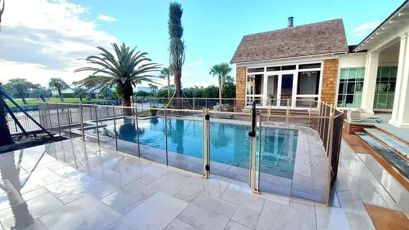 Luxury poolside patio with glass railing, palm trees, and a house beside the water under a blue sky