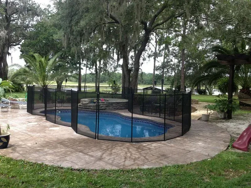 Backyard pool enclosed by black safety fencing, surrounded by trees and patio paving