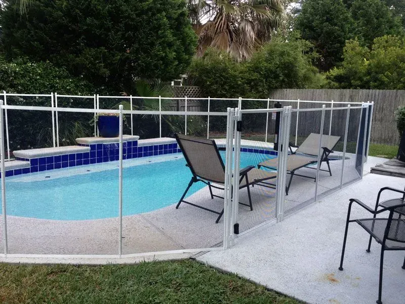 Backyard pool with safety fence, lounge chairs, and a blue spa beside trees