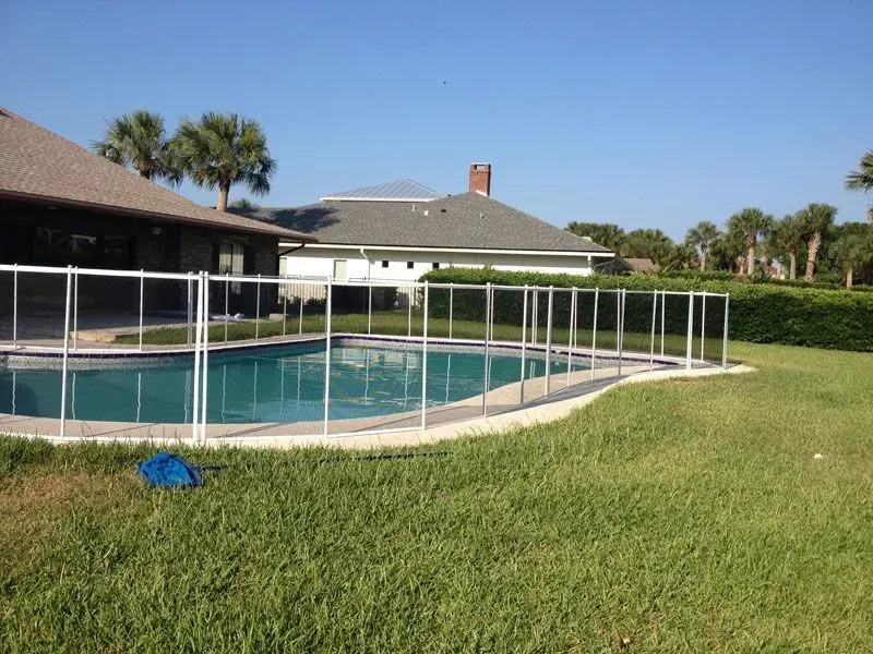 Backyard pool with white safety fence, green lawn, and a house under a clear blue sky