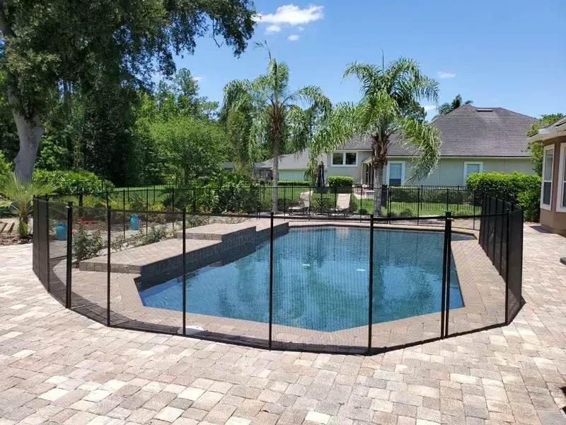 Backyard swimming pool enclosed by black safety fence, with patio and palm trees around it.