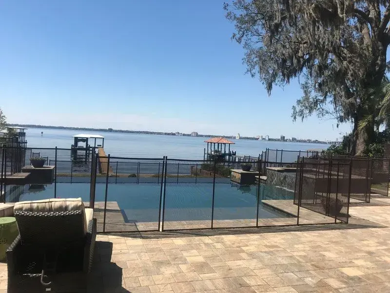 Poolside patio with a waterfront view, metal fence, and a tree under a clear blue sky.