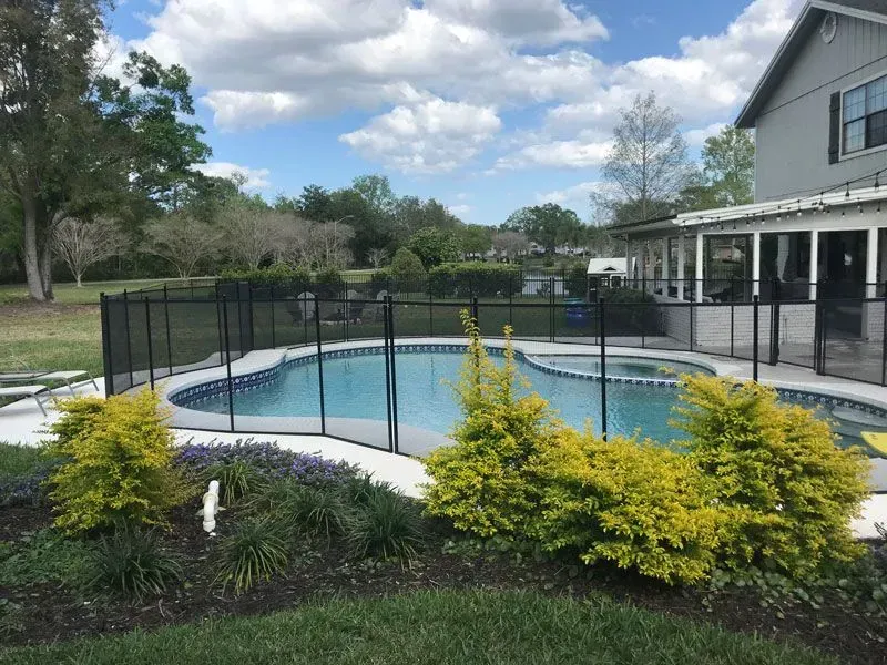 Backyard pool with black safety fence, yellow shrubs, and a house under a cloudy sky