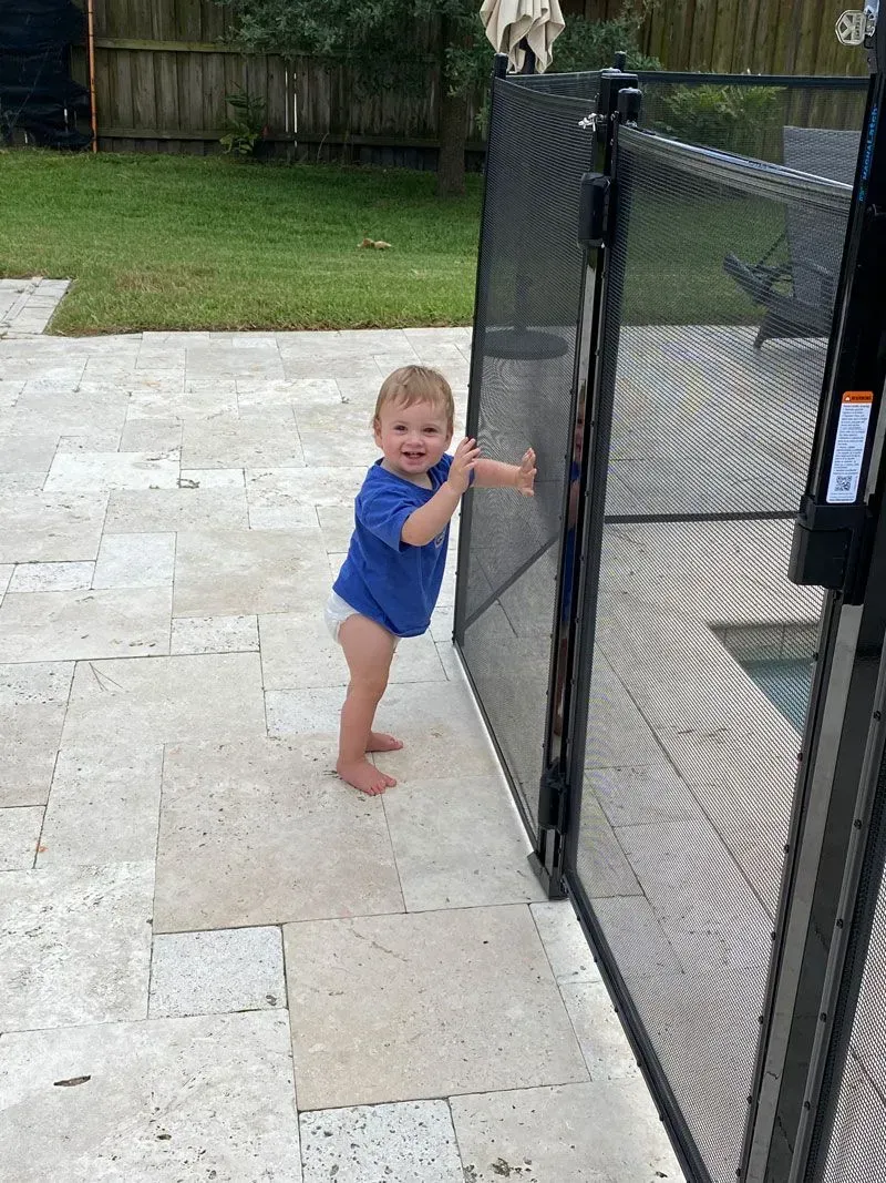 Toddler in a blue swimsuit holding a black gate beside a pool deck