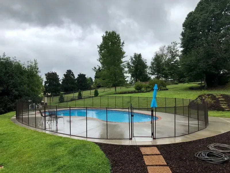 Backyard swimming pool enclosed by black safety fence, with blue umbrella and green lawn on a cloudy day