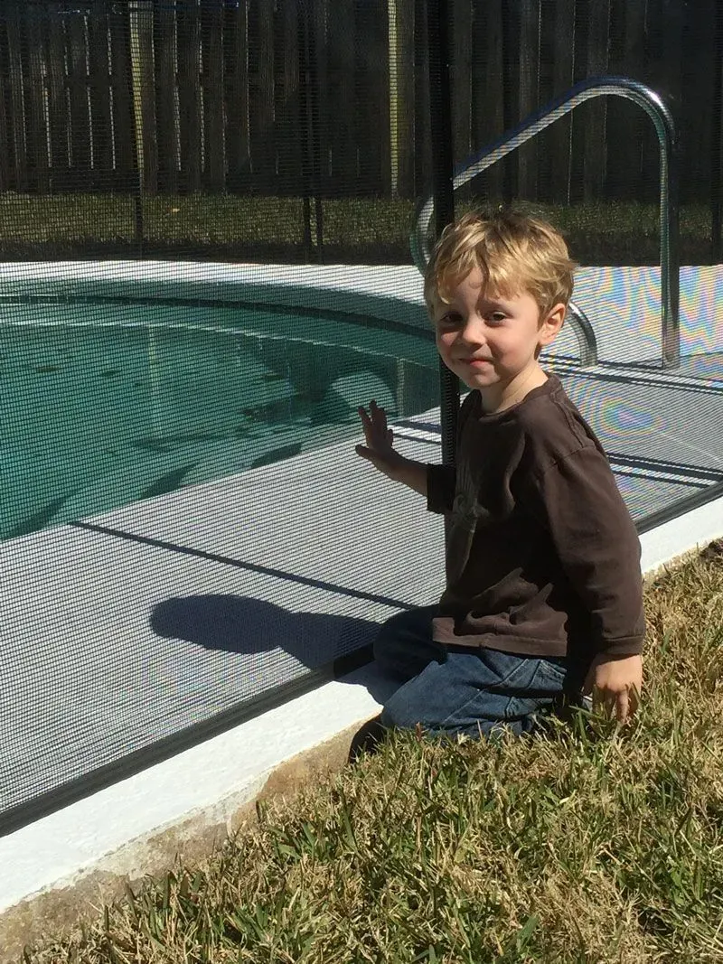 Child sitting by a backyard pool, holding the net fence and smiling at the camera.