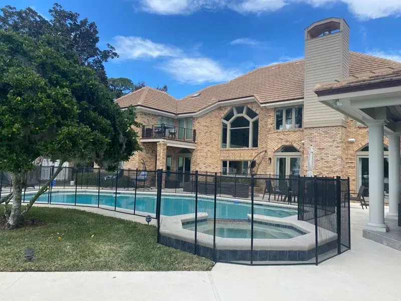 Backyard pool with safety fence beside a stone house under a blue sky