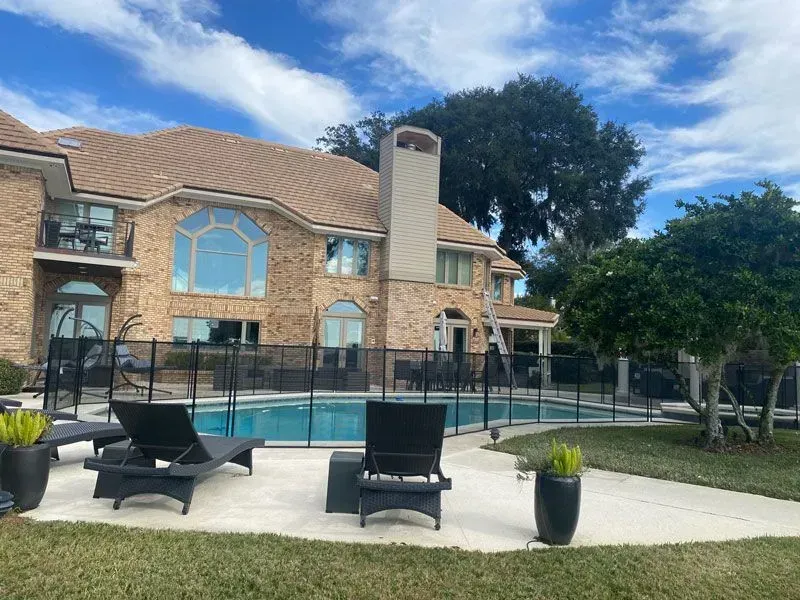 Backyard pool with patio chairs, glass fence, and a large stone house under a blue sky