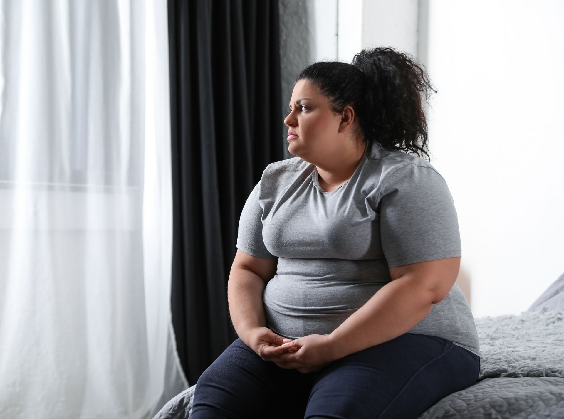 An obese white woman sitting on a bed looking depressed