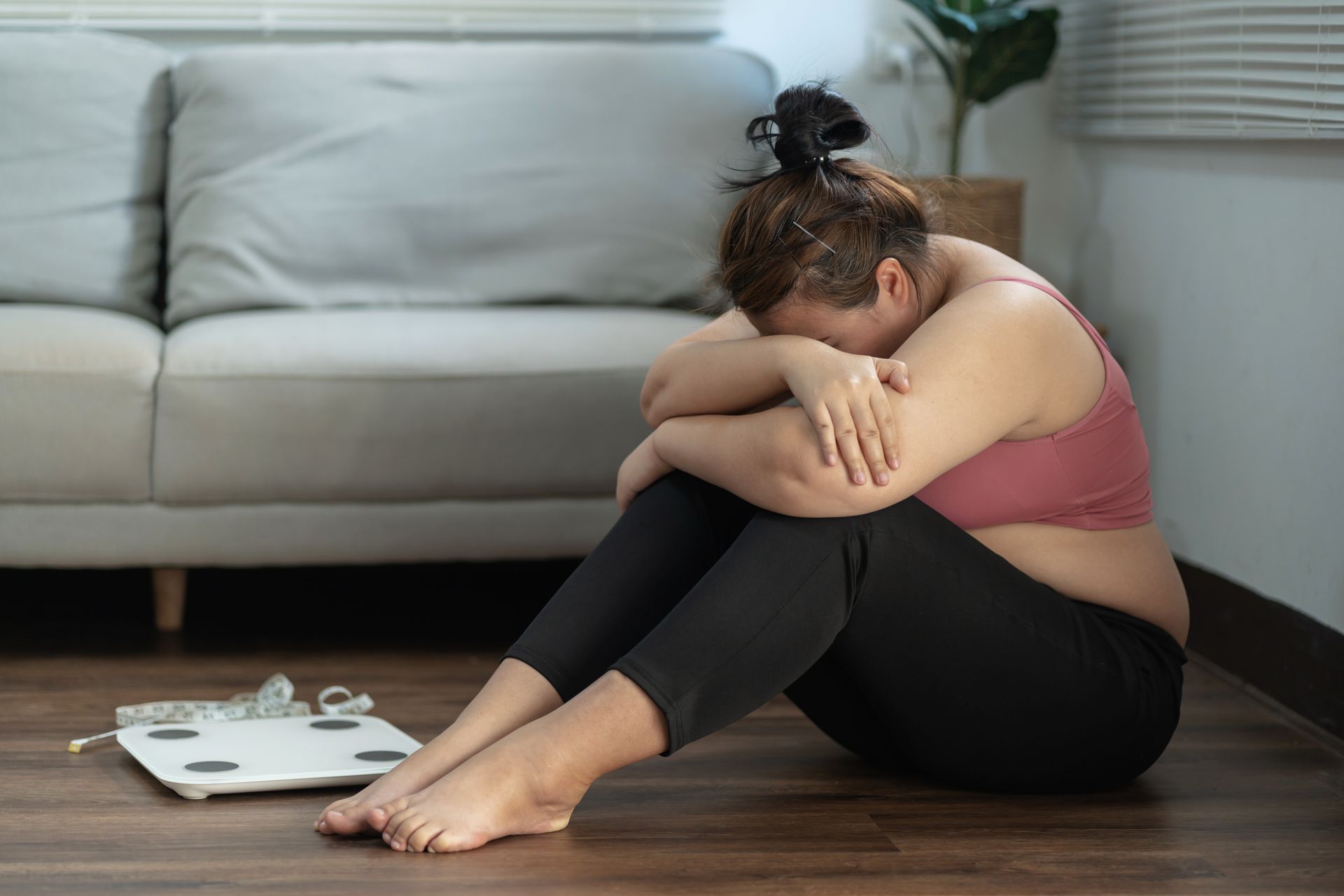 An overweight white woman sitting on the ground in front of a scale with her head in her arms depressed