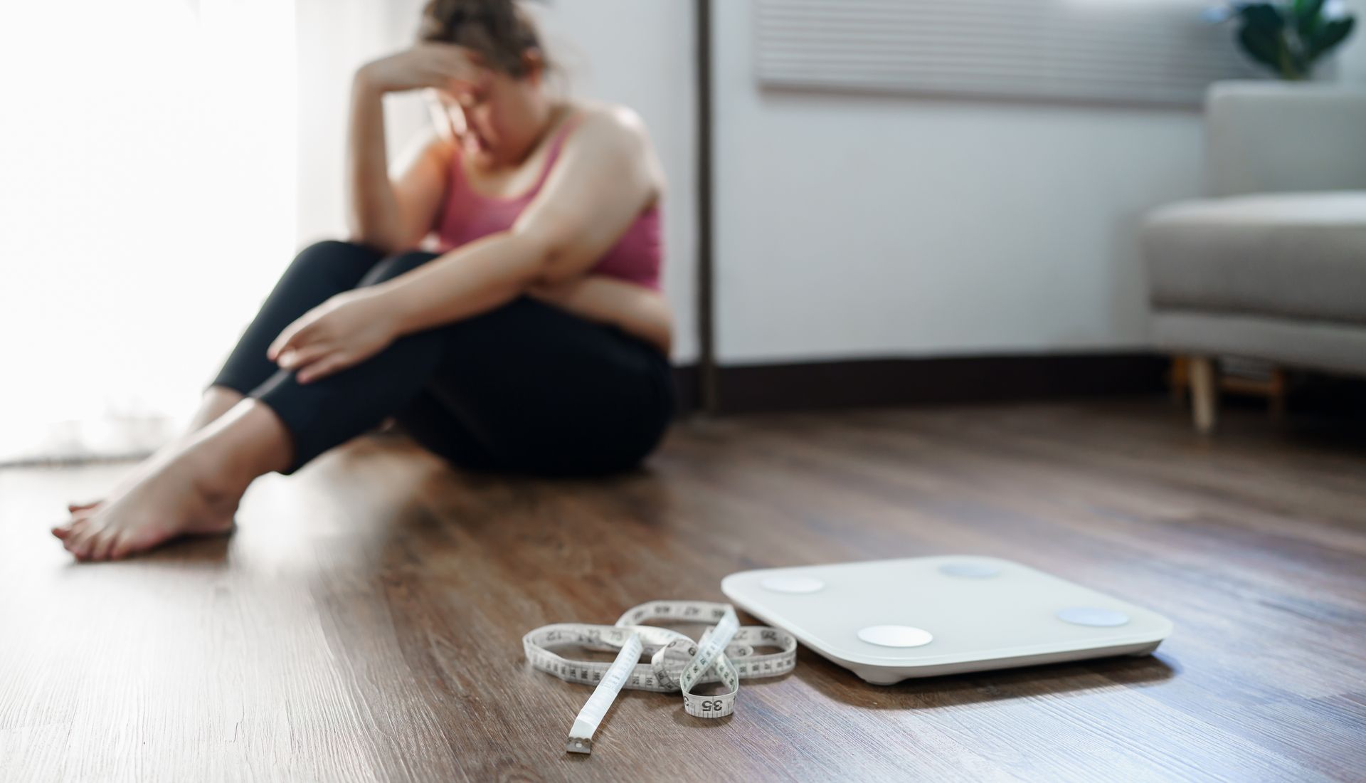 An obese white woman sitting on the floor next to a scale looking sad