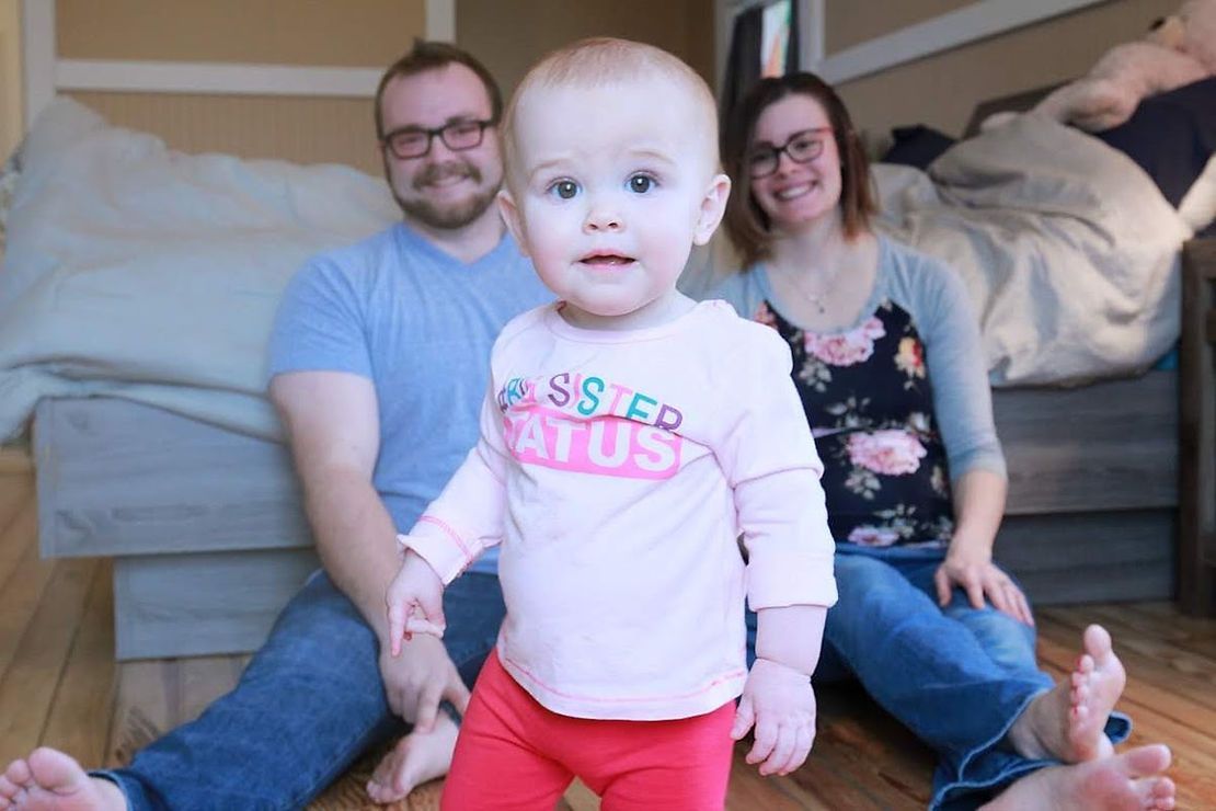 Family of three: a toddler in pink, a man in blue, and a woman in floral, on wooden floor.