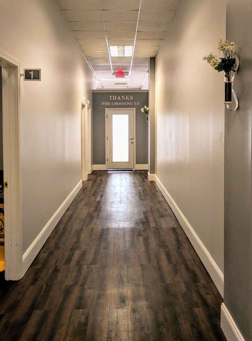 Narrow hallway with dark wood floor, white walls, and a bright door at the end.
