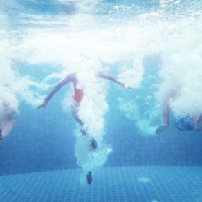 People underwater in a blue pool, arms outstretched with bubbles rising.