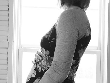Pregnant woman by window, holding belly, wearing floral dress and gray sweater.
