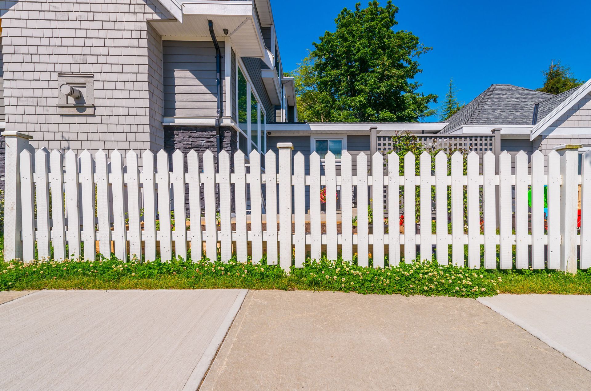 White picket fence in front of a gray house with a concrete sidewalk and green trees behind it