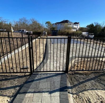 Black metal gate blocking a paved driveway under a clear blue sky, with houses in the background