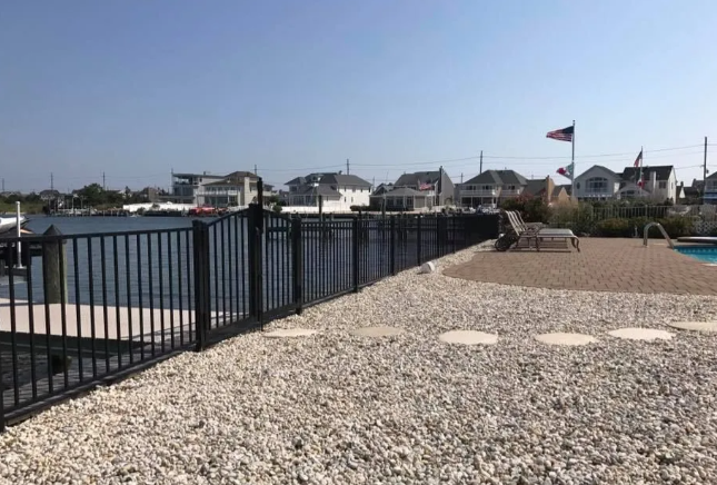 Riverside walkway with black railing, gravel ground, and houses across the water under a clear sky