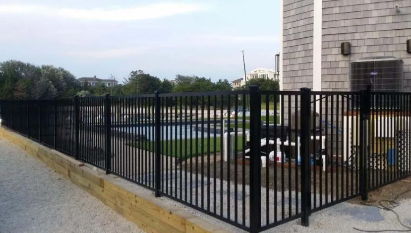 Black metal fence beside a beige building and parking lot on a gravel walkway.