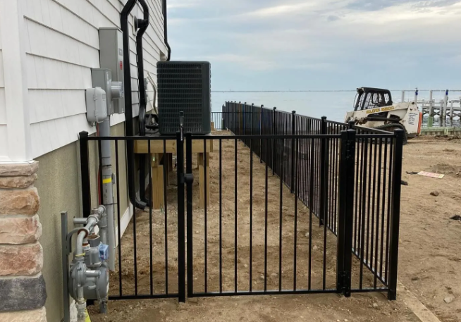 Black metal gate beside a house, closing off a narrow dirt walkway along the side yard.