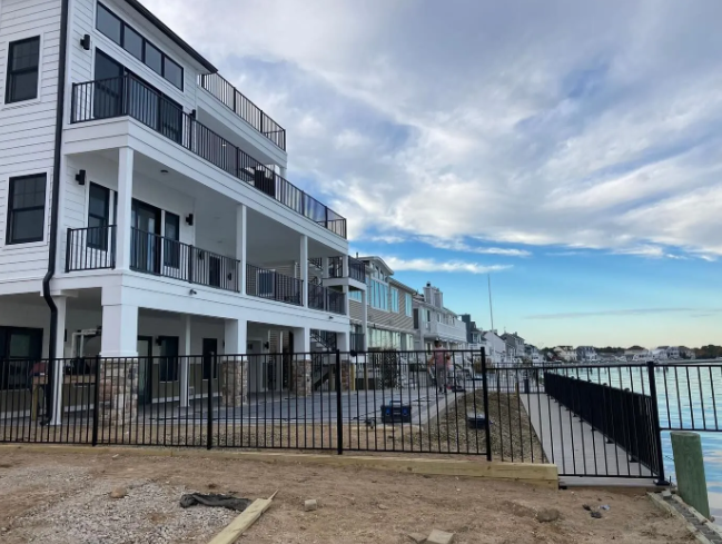 Row of modern waterfront condos with balconies beside a fenced shoreline under a cloudy sky